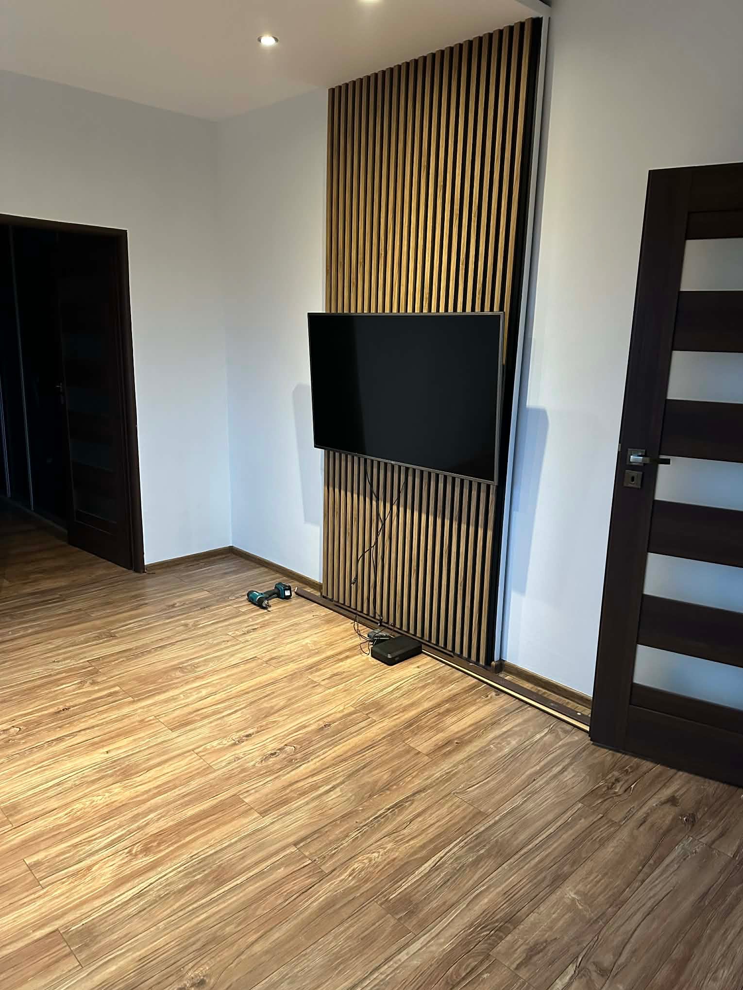 Nowoczesny salon po malowaniu: białe ściany, drewniane lamele za telewizorem, drewniana podłoga. W tle ciemne drzwi i wkrętarka na podłodze.