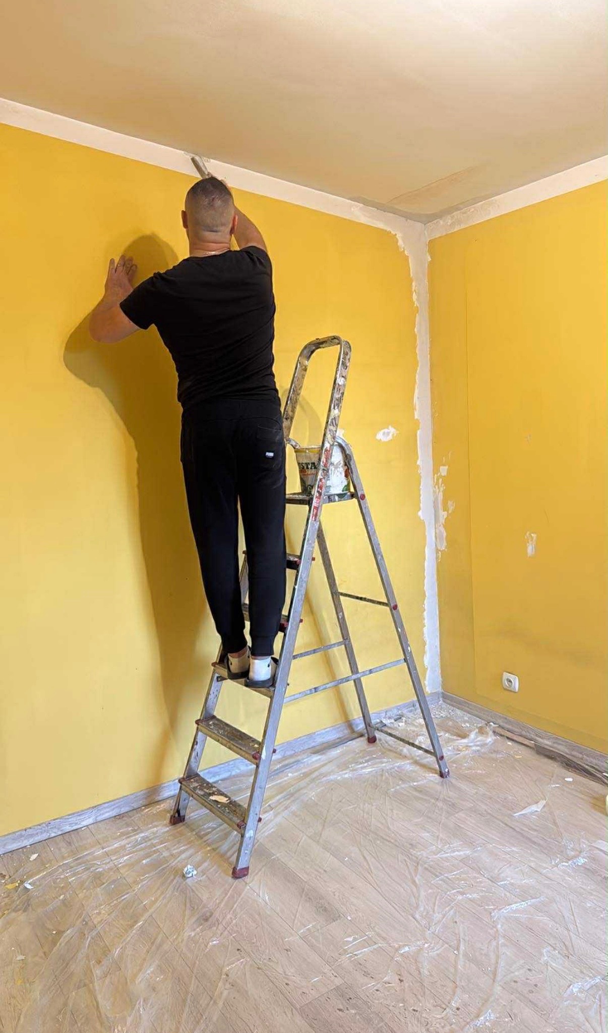 Mężczyzna na drabinie szpachluje żółtą ścianę przy suficie. Podłoga zabezpieczona folią. Widoczny fragment narożnika i puszka z farbą.