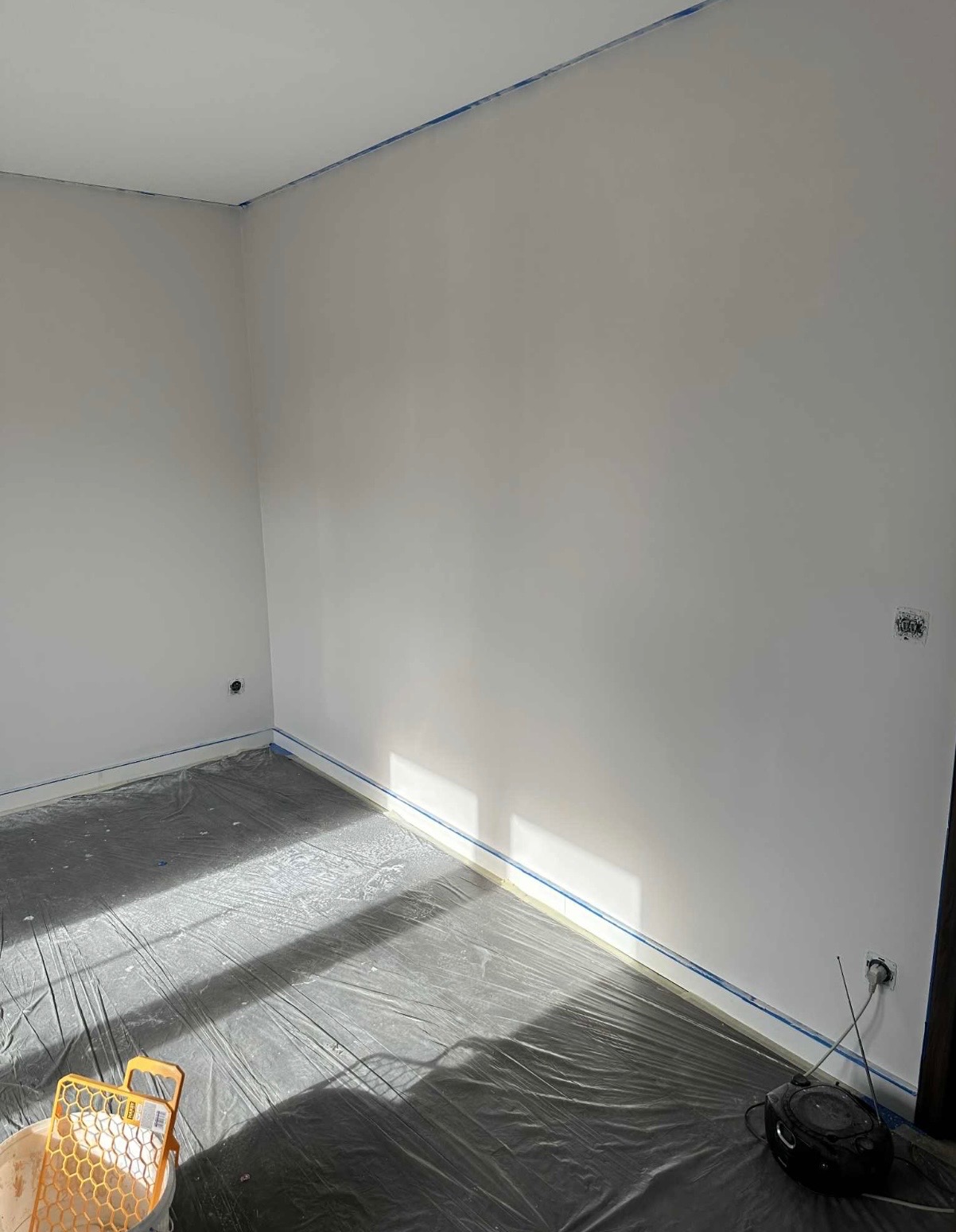 Świeżo pomalowane na biało wnętrze pokoju, zabezpieczone folią i taśmą malarską, z widocznym wiaderkiem i radiem. Promienie słoneczne podkreślają gładkość ścian.