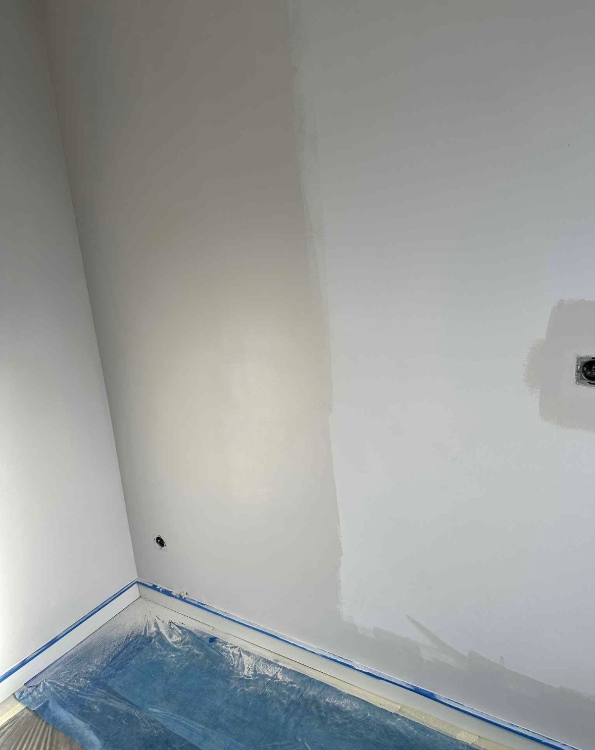 Świeżo pomalowany, jasnoszary róg pokoju z zabezpieczoną folią podłogą i taśmą malarską. Widoczny fragment ściany z gniazdkiem elektrycznym.