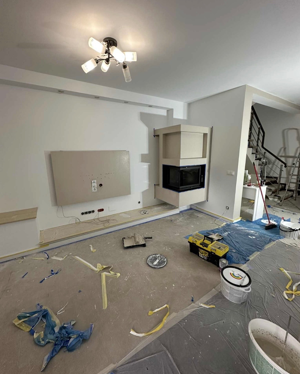 Wnętrze w trakcie odświeżania: świeżo pomalowane ściany na biało, zabezpieczona podłoga folią i taśmą malarską, widoczny kominek i narzędzia malarskie.