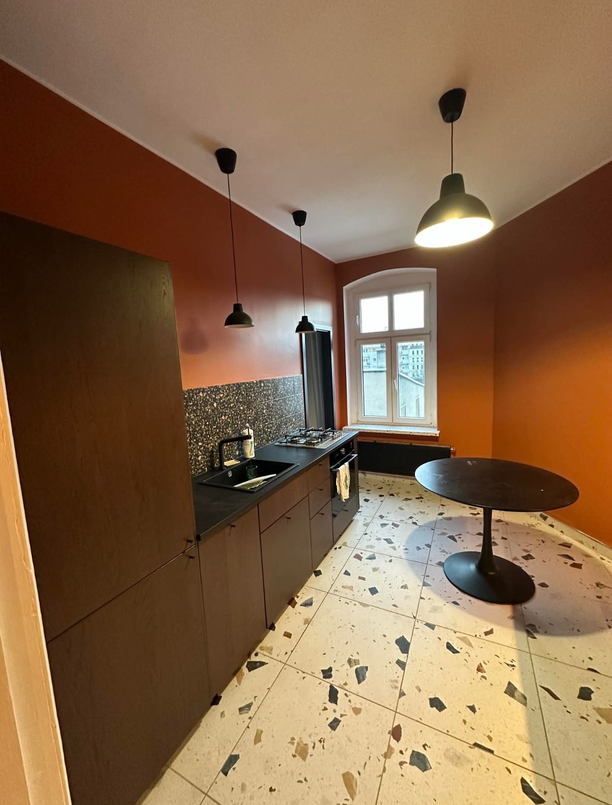 Kuchnia po remoncie: ściany w odcieniach terakoty, ciemne meble kuchenne z czarnym blatem, podłoga w stylu lastryko. Widok na okno i stół.