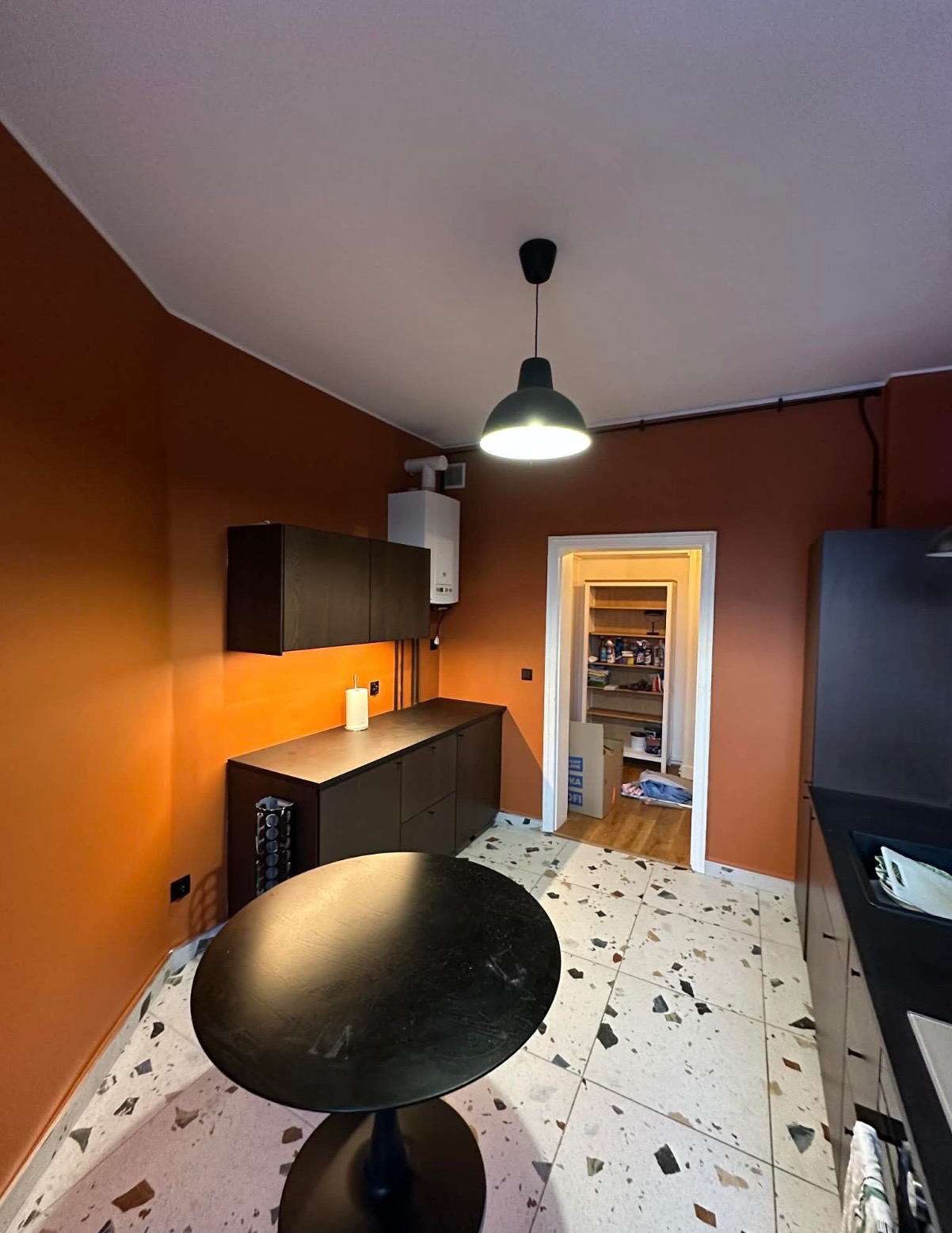 Pomarańczowa kuchnia po malowaniu: ciemne szafki, okrągły stół i wzorzysta podłoga. Widoczne rury i piec gazowy. Oświetlenie: czarna lampa sufitowa.