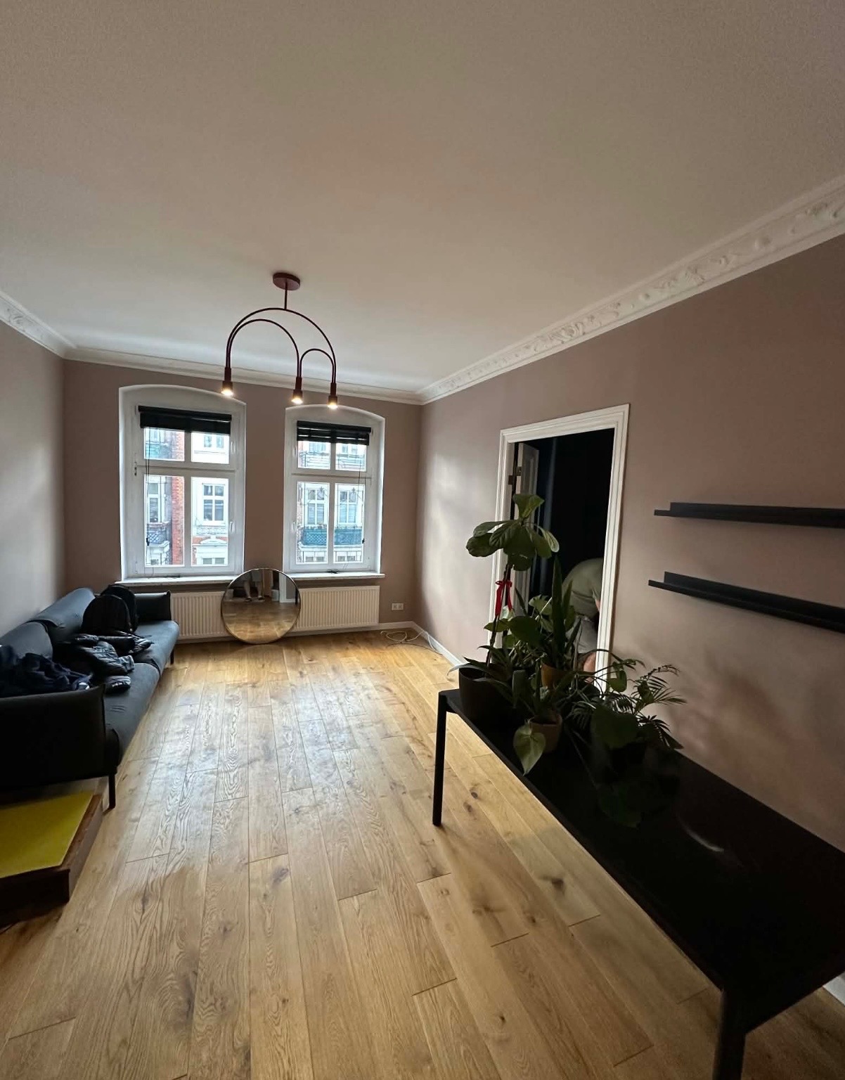 Jasne wnętrze po malowaniu: ściany w odcieniach beżu, drewniana podłoga, nowoczesne oświetlenie i dekoracyjne listwy sufitowe. Widok na okno i rośliny doniczkowe.