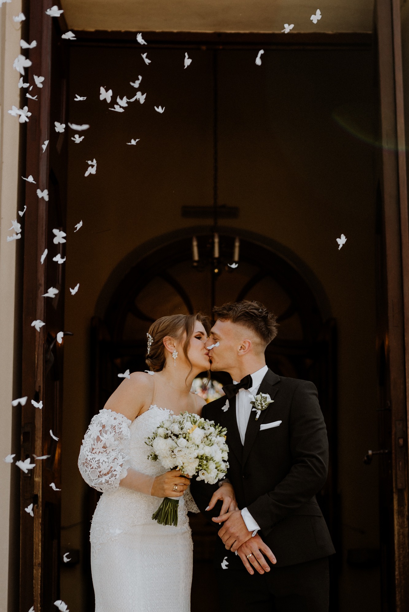 Pocałunek nowożeńców przed kościołem, otoczonych białymi konfetti w kształcie motyli. Panna młoda w koronkowej sukni, pan młody w czarnym smokingu z muchą.