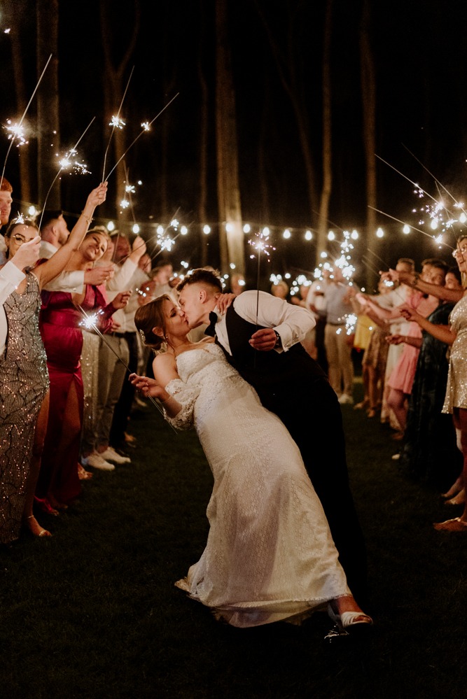 Pocałunek nowożeńców w otoczeniu gości z zimnymi ogniami, nocna sceneria z drzewami i girlandami świateł. Panna młoda w koronkowej sukni, pan młody w czarnym garniturze.