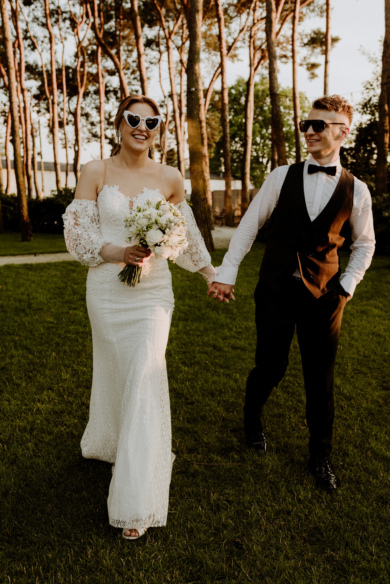 Uśmiechnięta para młoda w okularach w kształcie serc spaceruje po zielonym trawniku, trzymając się za ręce na tle drzew w słoneczny dzień. Romantyczna sesja ślubna.