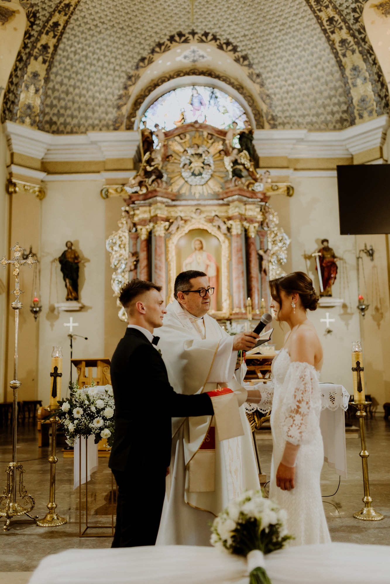 Uroczystość ślubna w kościele: para młoda, ksiądz z mikrofonem, bogato zdobione wnętrze z ołtarzem i witrażami. Ciepłe, naturalne światło podkreśla sakralny charakter.