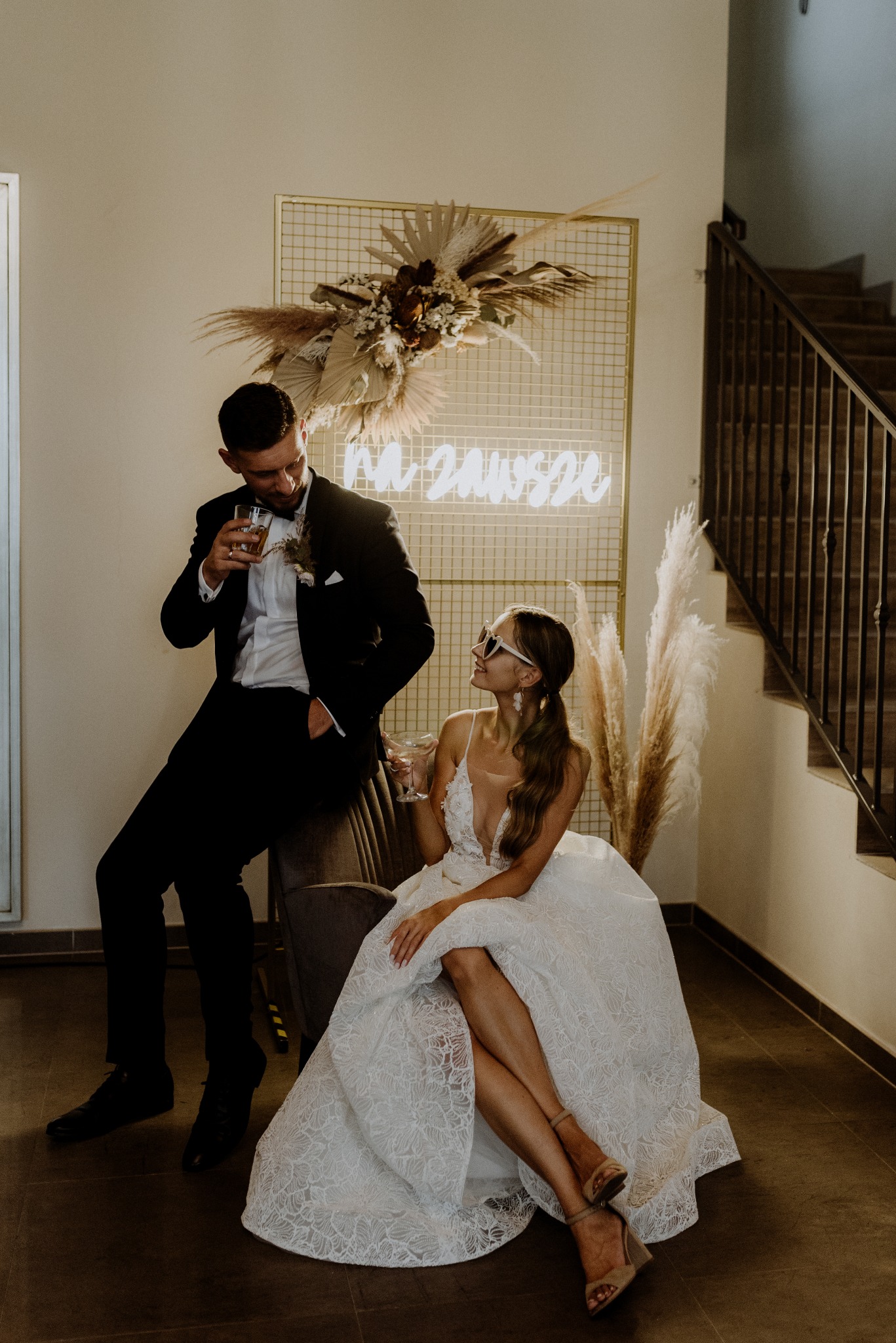 Elegancka para młoda w stylizacji boho, on w smokingu z drinkiem, ona w sukni z koronką i okularach, siedząca na fotelu, w tle dekoracja z neonem 'Na zawsze'.