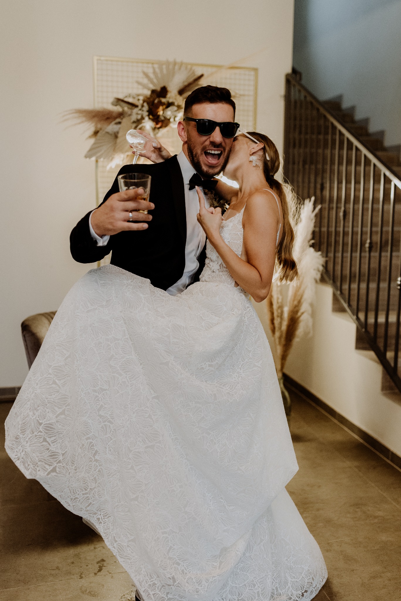 Rozbawiony pan młody w okularach i z drinkiem, podnoszący pannę młodą w koronkowej sukni. W tle schody i dekoracje z suszonych traw.
