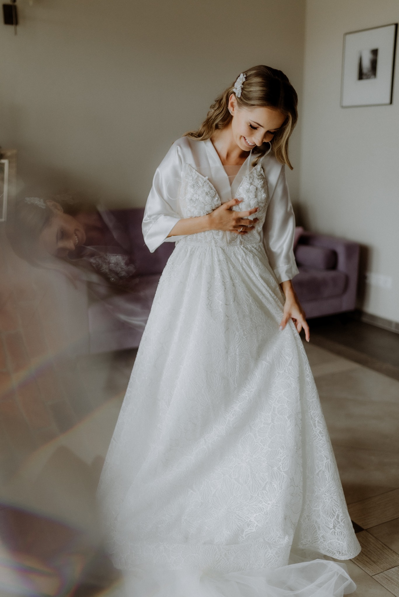 Uśmiechnięta panna młoda w satynowym szlafroku, trzymająca suknię ślubną z koronkowymi zdobieniami, w jasnym wnętrzu z fioletową sofą w tle.