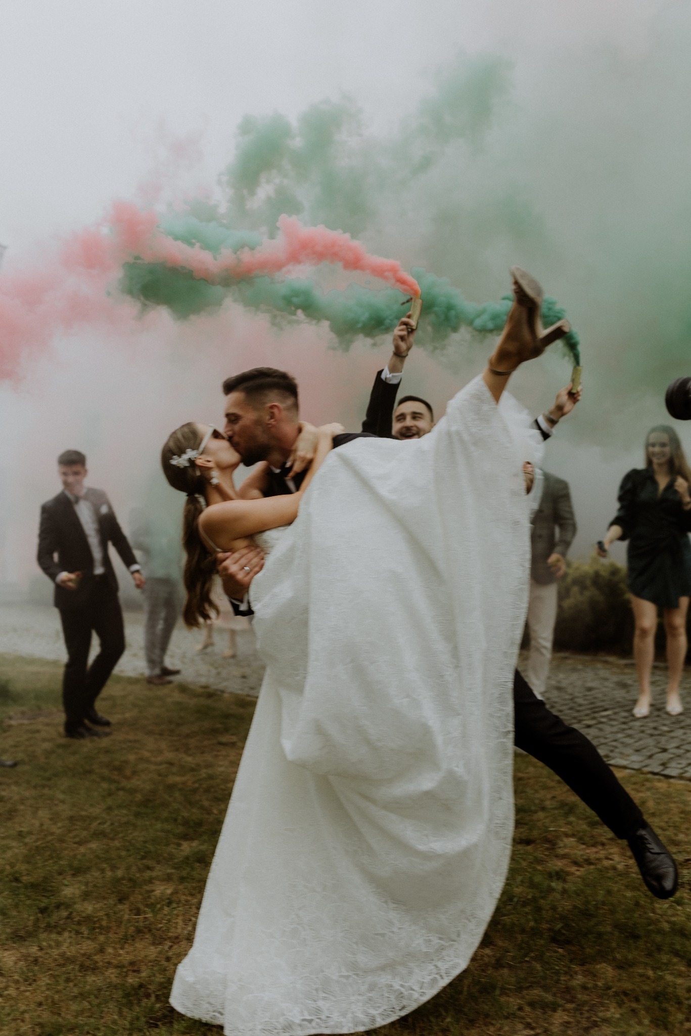 Panna młoda w białej sukni ślubnej, unoszona przez pana młodego podczas pocałunku, w tle kolorowe dymy i goście weselni. Ujęcie z dołu, dynamiczne.