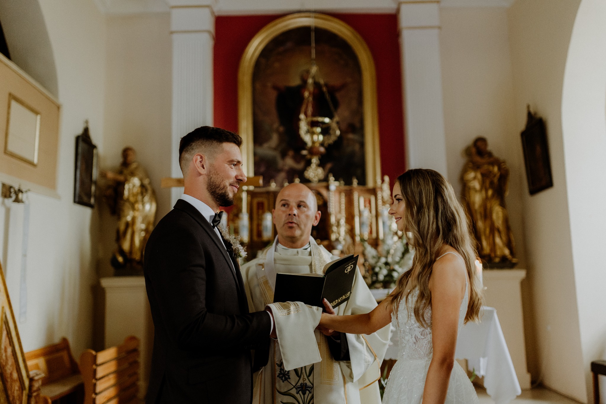 Uroczystość ślubna w kościele: para młoda w eleganckich strojach, ksiądz z księgą, złote figury w tle. Ciepłe, naturalne światło podkreśla sakralny charakter.