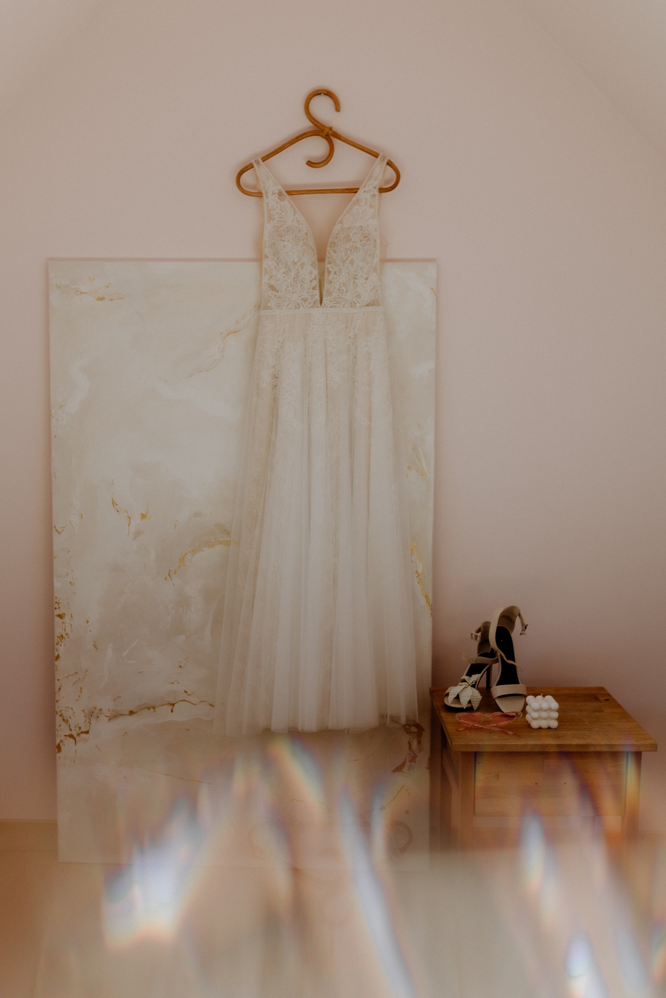 Delikatna suknia ślubna z koronkową górą, wisząca na wieszaku obok marmurowej płyty. Na stoliku obok leżą buty na obcasie i dekoracyjna świeca.