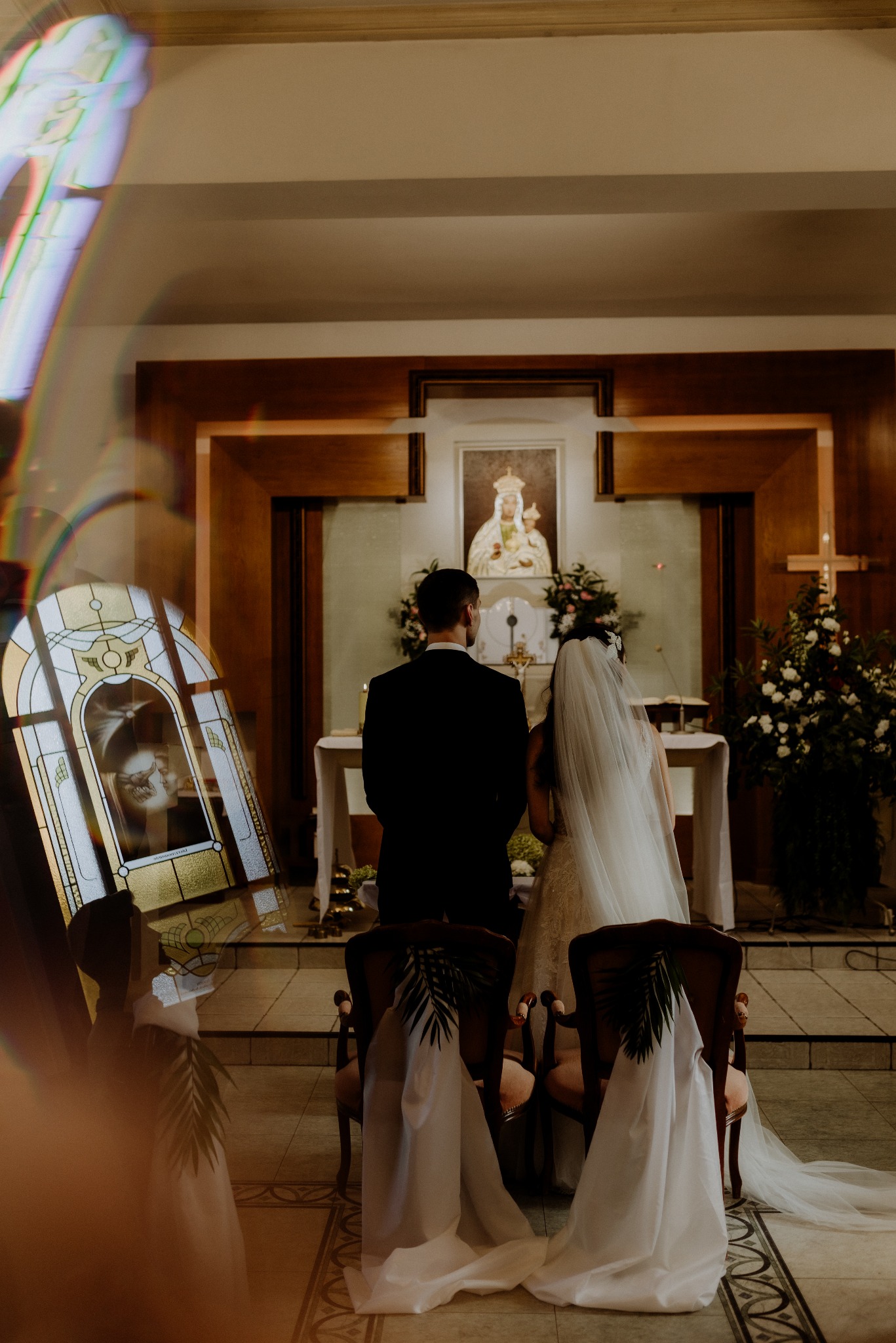 Para młoda w kościele, widziana od tyłu, z dekoracjami kwiatowymi i witrażem odbijającym ich wizerunek. Ujęcie z delikatnym efektem flary obiektywu.