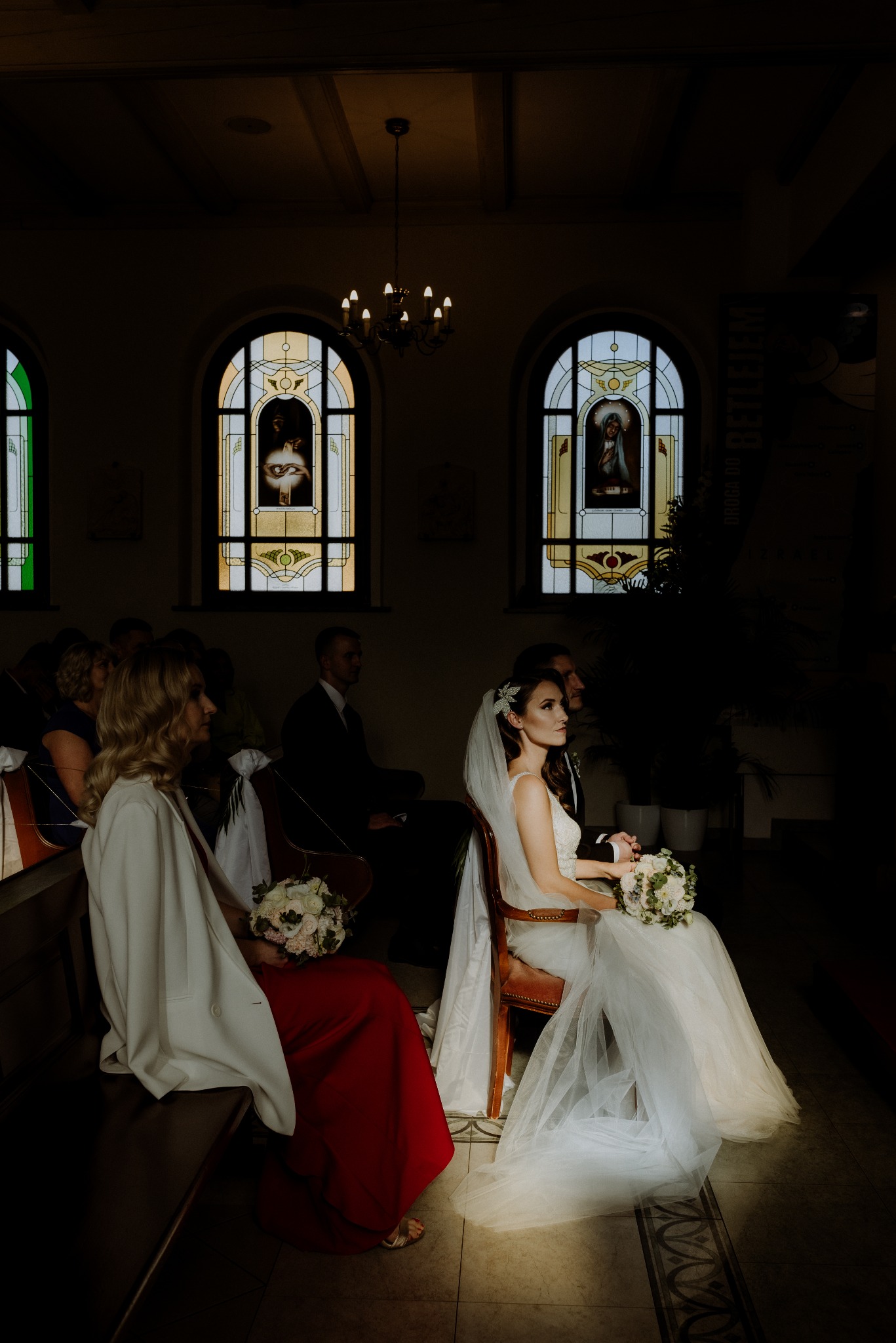 Panna młoda w kościele, w jasnej sukni i welonie, siedzi na krześle z bukietem. W tle witraże i żyrandol, a obok goście weselni. Ciemne wnętrze kościoła.