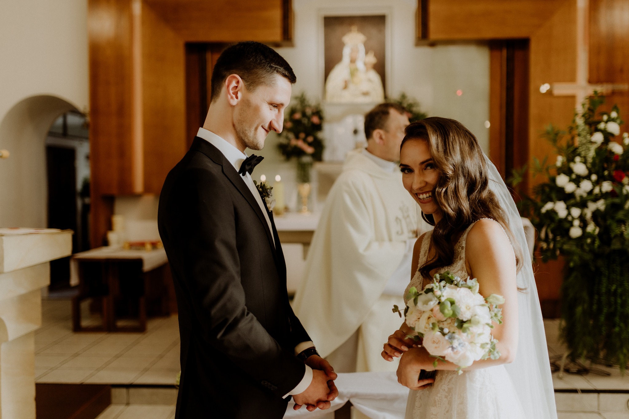 Uśmiechnięta panna młoda z bukietem i elegancki pan młody podczas ceremonii ślubnej w kościele, w tle ksiądz i dekoracje kwiatowe.