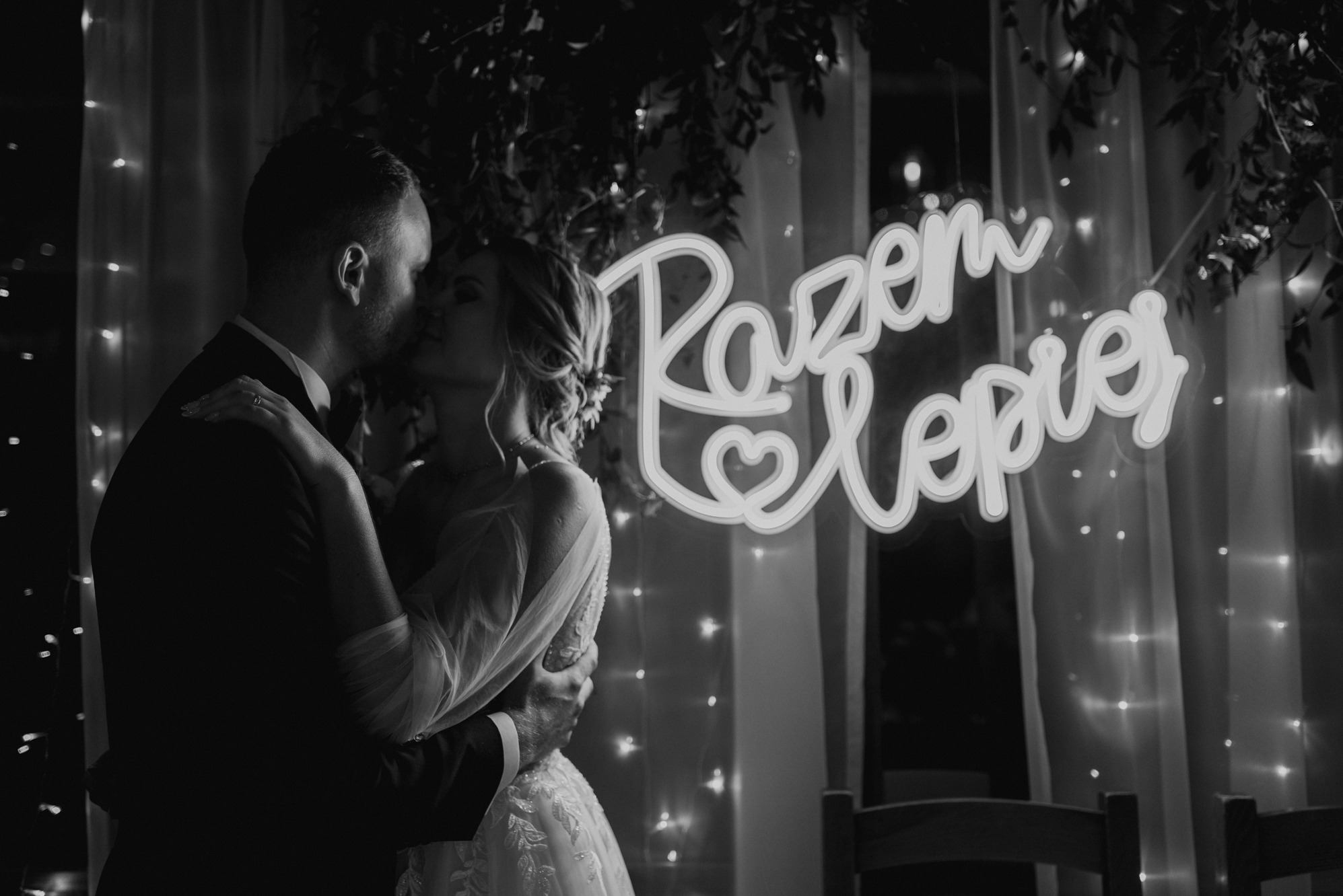 Czarno-białe zdjęcie pary młodej w objęciach, z neonowym napisem 'Razem Lepiej' w tle, oświetlonymi girlandami i delikatną dekoracją roślinną.