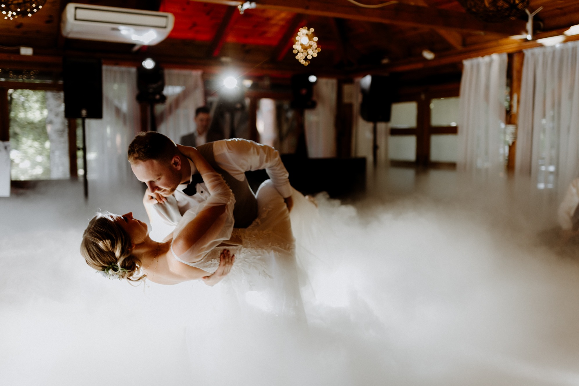 Para młoda w tańcu, otoczona mgłą na parkiecie. Pan młody w smokingu, panna młoda w białej sukni. W tle drewniana konstrukcja sali i oświetlenie.