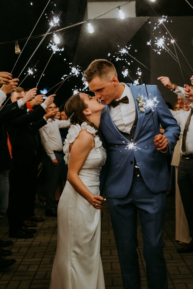 Pocałunek pary młodej w Siechnicach, otoczonej gośćmi z zimnymi ogniami. Panna młoda w białej sukni, pan młody w niebieskim garniturze. Romantyczna atmosfera.