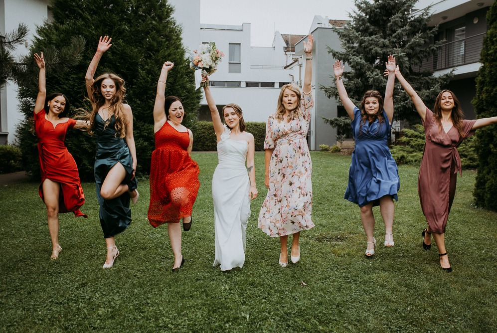 Radosne druhny w kolorowych sukienkach skaczą na trawie, świętując z panną młodą trzymającą bukiet. Uśmiechy, energia i pozytywna atmosfera w plenerze.
