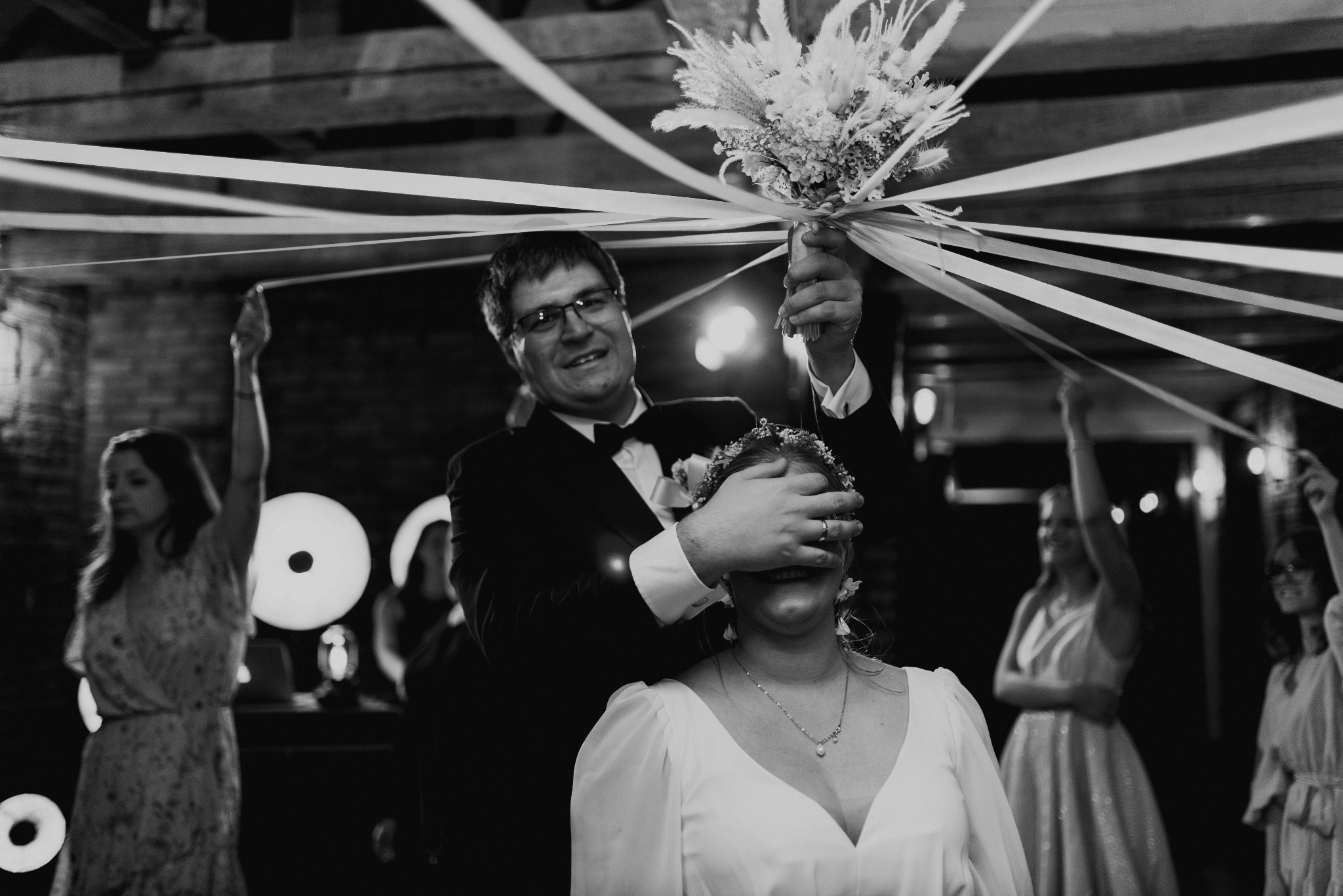Czarno-białe zdjęcie z wesela: Pan młody zasłania oczy pannie młodej, trzymając bukiet nad jej głową. W tle goście z podniesionymi wstążkami.
