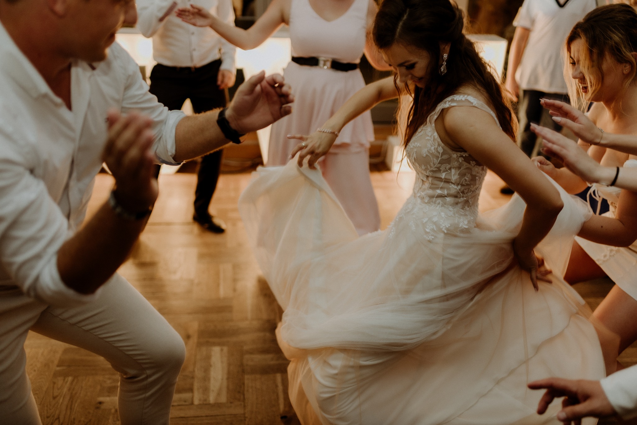 Uśmiechnięta panna młoda w koronkowej sukni tańczy na parkiecie, otoczona gośćmi weselnymi. Dynamiczne ujęcie z bliska, ciepłe oświetlenie.