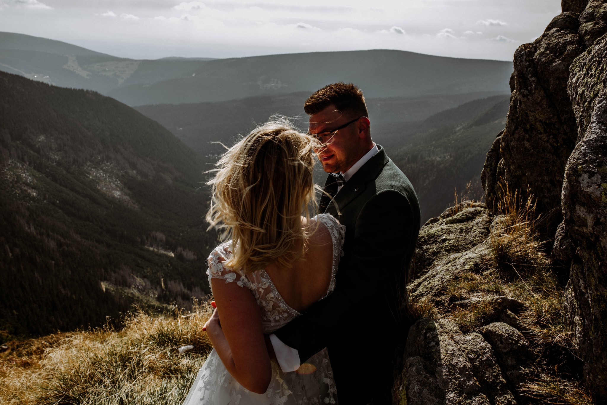 Para młoda w objęciach na tle górskiego krajobrazu. Panna młoda w koronkowej sukni, pan młody w ciemnym garniturze. Wietrzna pogoda, naturalne światło.