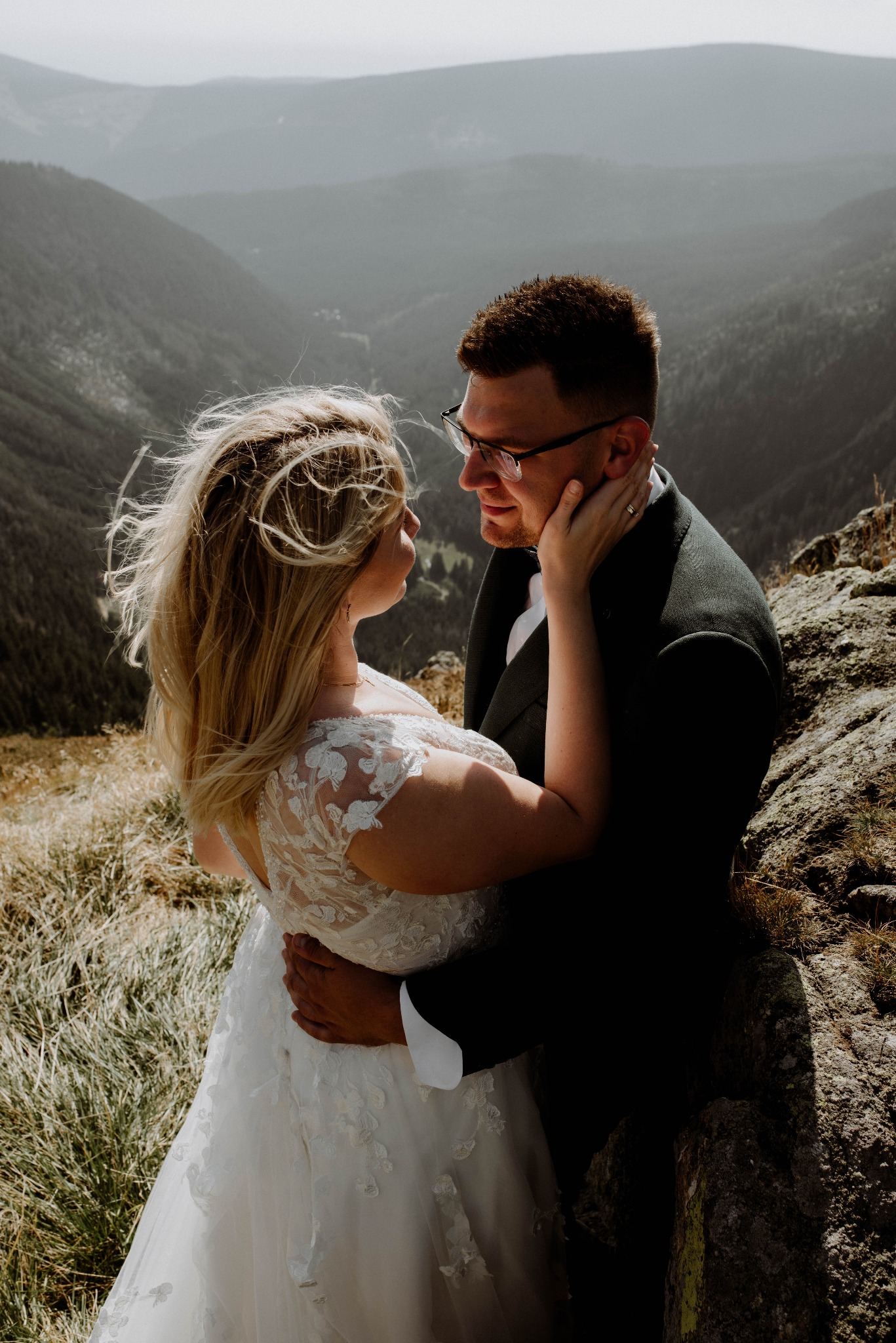 Para młoda w objęciach na tle górskiego krajobrazu. Panna młoda w koronkowej sukni, pan młody w ciemnym garniturze. Romantyczna sesja plenerowa w naturalnym świetle.