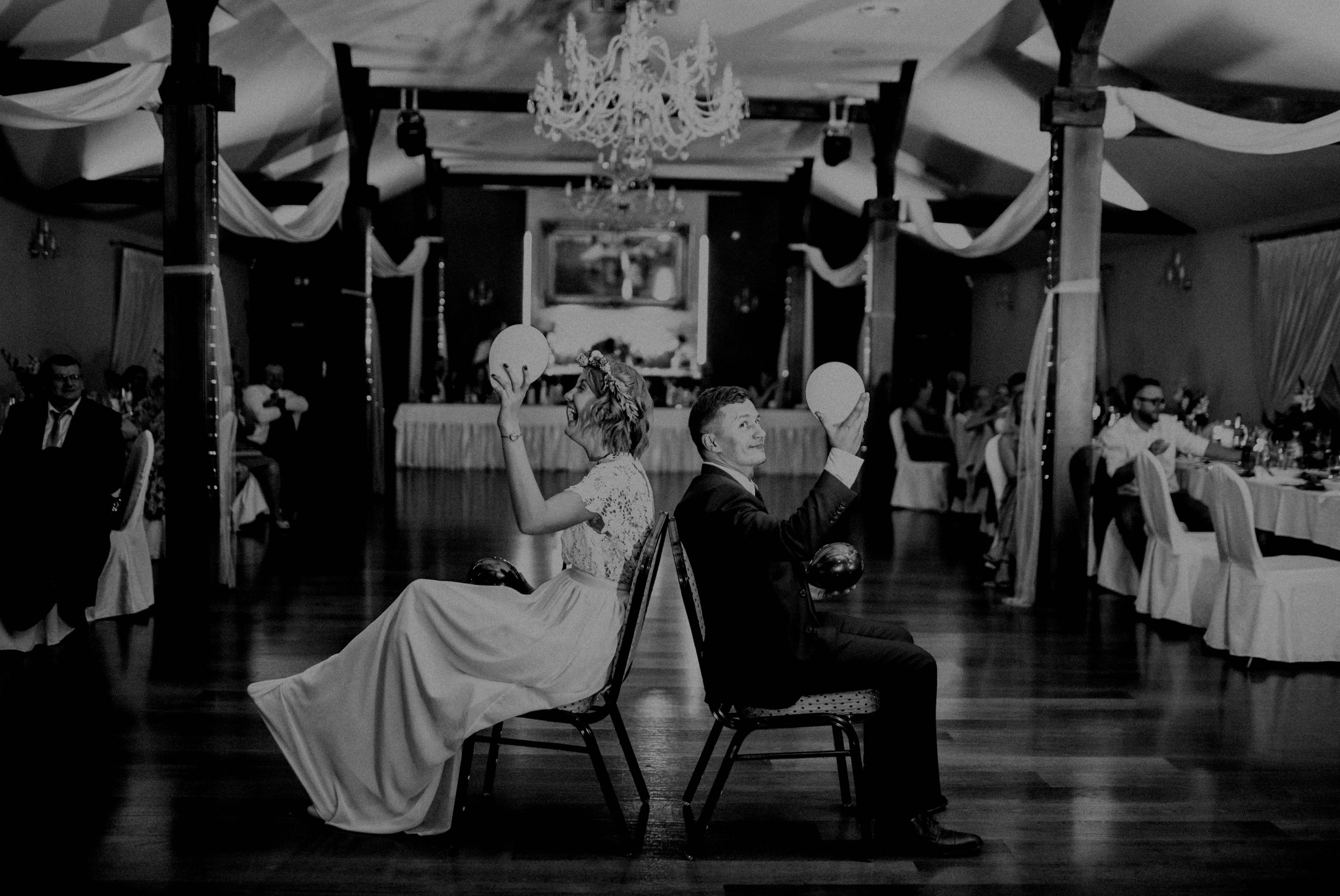 Czarno-białe zdjęcie pary młodej siedzącej tyłem do siebie na krzesłach, trzymającej balony na sali weselnej z żyrandolem i gośćmi w tle. Nietypowe ujęcie.