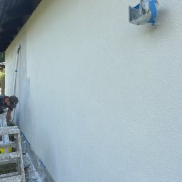 OŻIBUD Andrzej Ożóg - Malowanie elewacji domu: mężczyzna na drabinie maluje ścianę na biało, zabezpieczone elementy, teksturowana powierzchnia, widok z boku, częściowo pomalowana ściana.