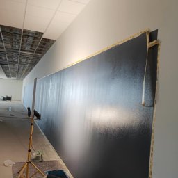 OŻIBUD Andrzej Ożóg - Ściana pomalowana na ciemny kolor w biurze, widoczne narzędzia malarskie, taśma malarska zabezpieczająca krawędzie, oświetlenie robocze. Prace wykończeniowe w toku.