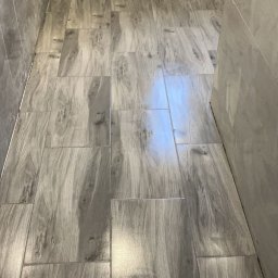 Ozibud - Ukończona łazienka z szarymi płytkami imitującymi drewno na podłodze i ścianach. Widoczne metalowe elementy instalacji wodnej.