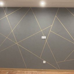 OŻIBUD Andrzej Ożóg - Szara ściana z geometrycznym wzorem z złotych linii, widoczne gniazdka elektryczne. Podłoga drewniana, jasne oświetlenie sufitowe.