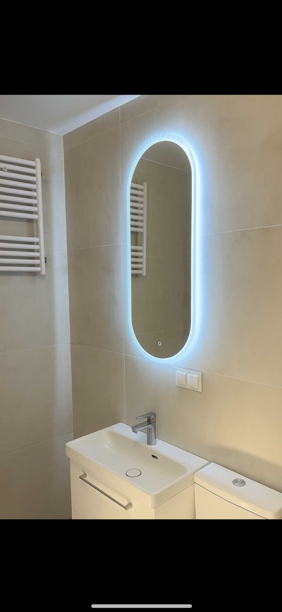 Nowoczesna łazienka z owalnym lustrem LED, białą umywalką z szafką i grzejnikiem drabinkowym. Beżowe płytki na ścianach. Minimalistyczny design.