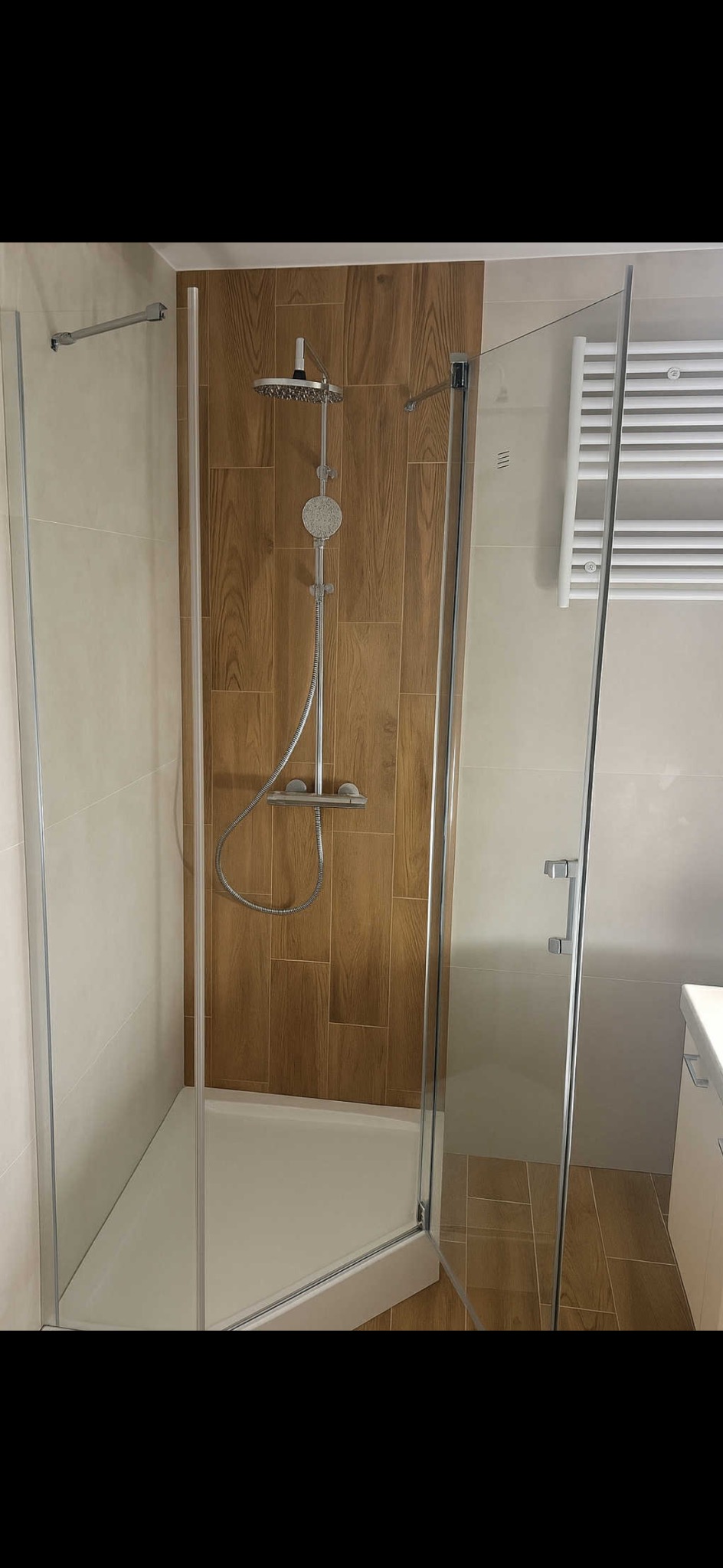 Kabina prysznicowa z deszczownicą, wykończona płytkami imitującymi drewno. Widoczny grzejnik drabinkowy i fragment umywalki z szafką. Nowoczesny design łazienki.