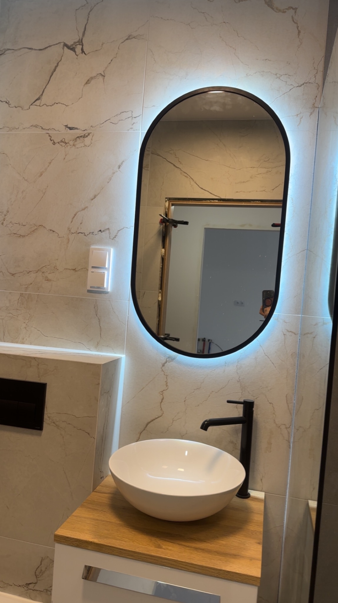 Nowoczesna łazienka z owalnym lustrem LED, białą umywalką na drewnianym blacie i marmurowymi płytkami. Widoczne detale wykończenia i oświetlenie.