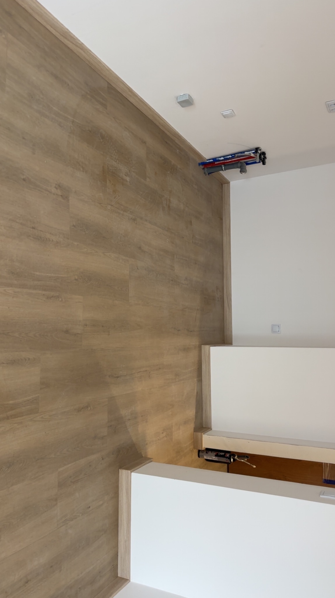 Jasne panele podłogowe ułożone wzdłuż ściany, biała ściana z instalacją elektryczną pod sufitem i fragment zabudowy meblowej. Ujęcie z góry.