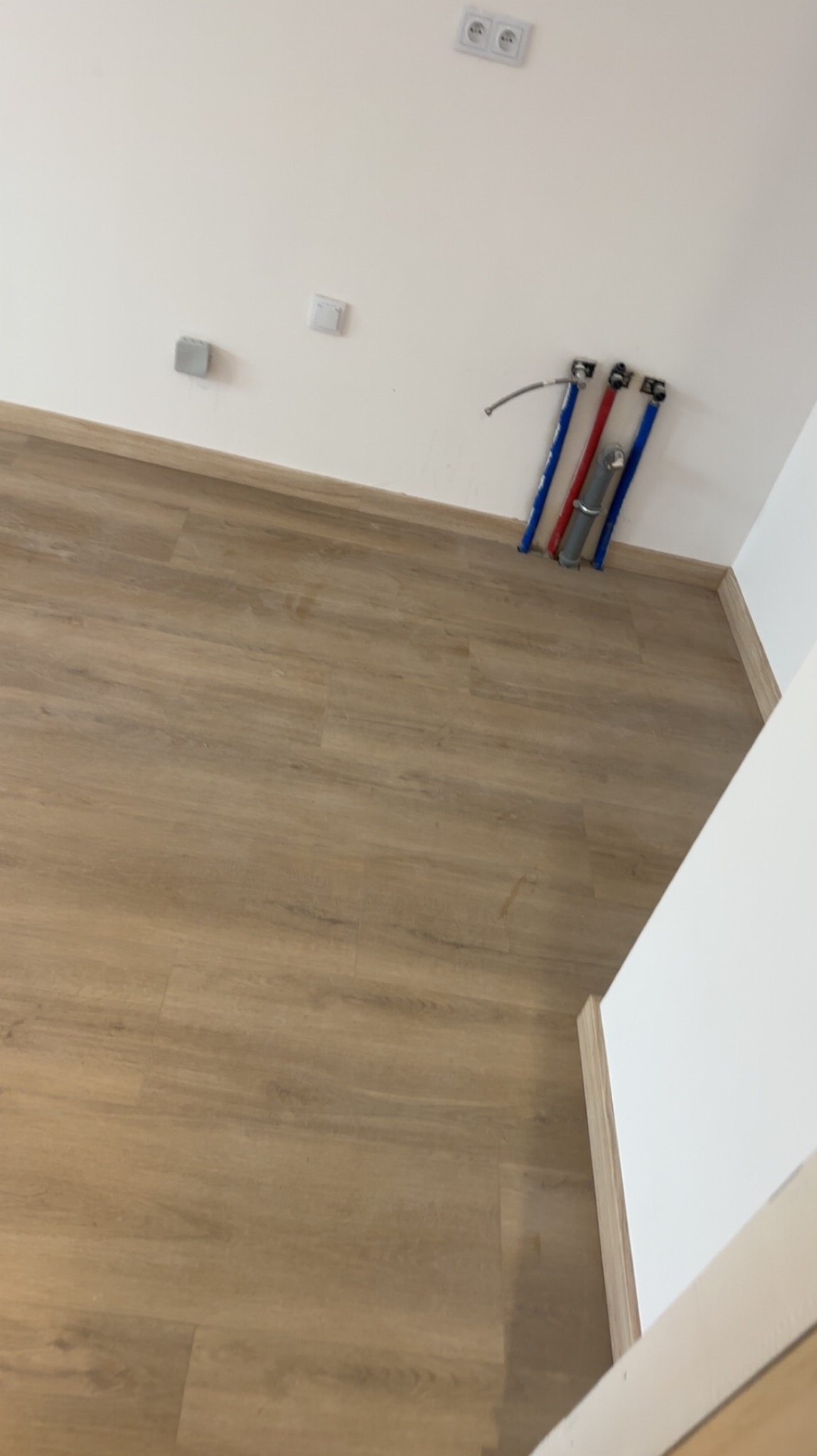 Jasne panele podłogowe w nowym wnętrzu, z widocznymi rurami instalacji wodnej przy ścianie. Minimalistyczny design, świeżo po remoncie, z gładkimi, białymi ścianami.