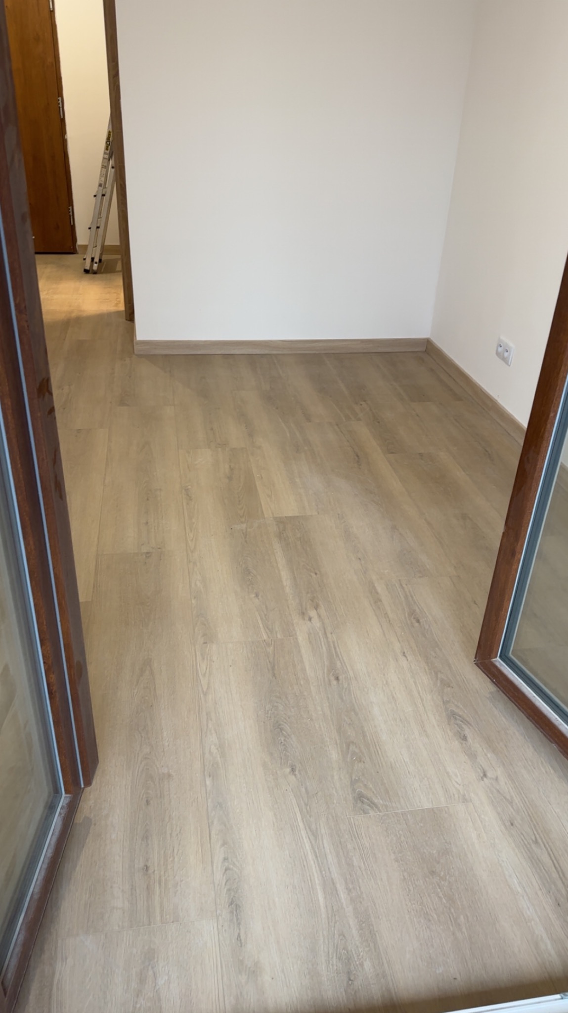 Jasne panele podłogowe ułożone w pustym pokoju z białymi ścianami i drewnianymi listwami przypodłogowymi. W tle drabina.