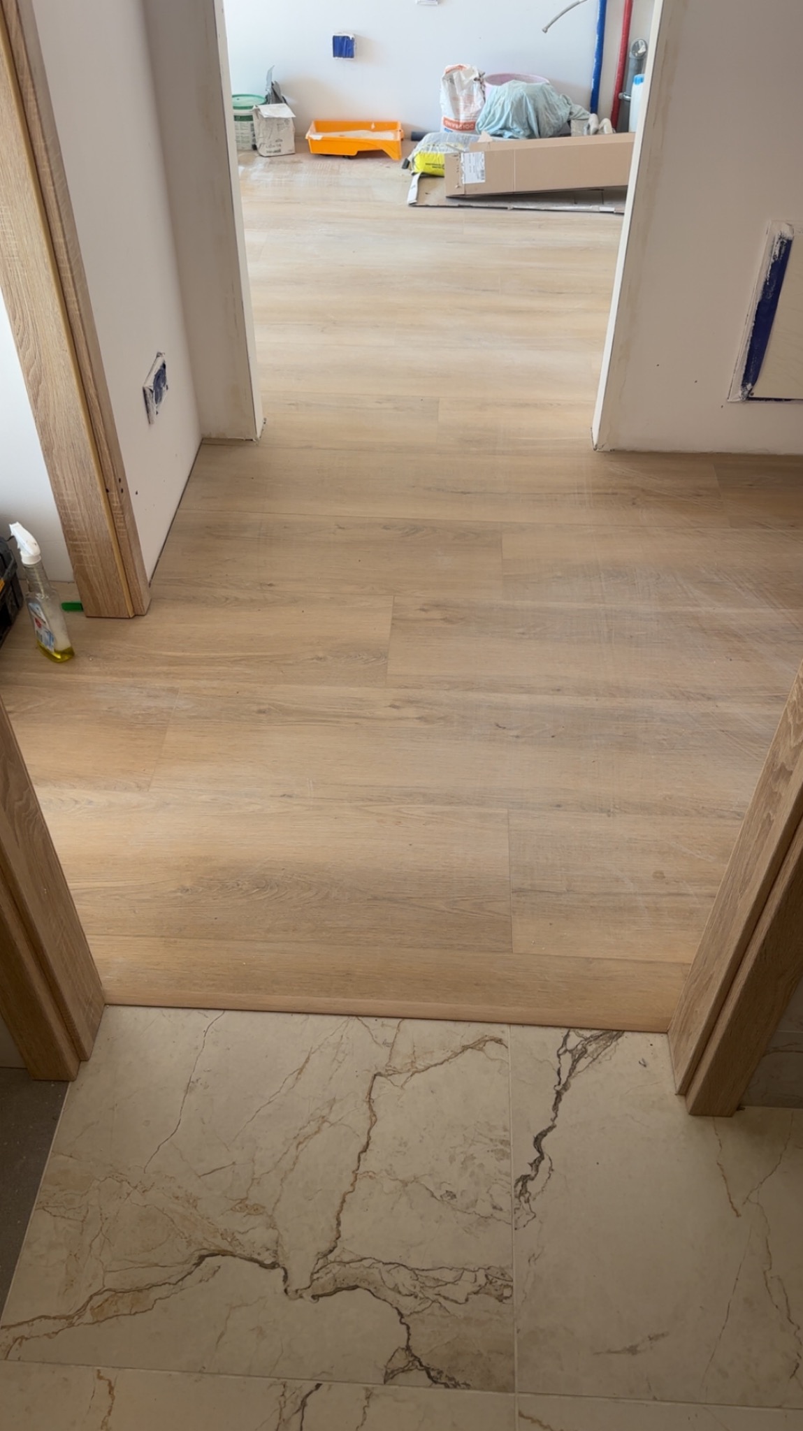 Ułożona podłoga z paneli w odcieniach beżu, widok z progu na marmurowe płytki. Częściowo widoczne materiały budowlane w tle sugerują trwający remont.