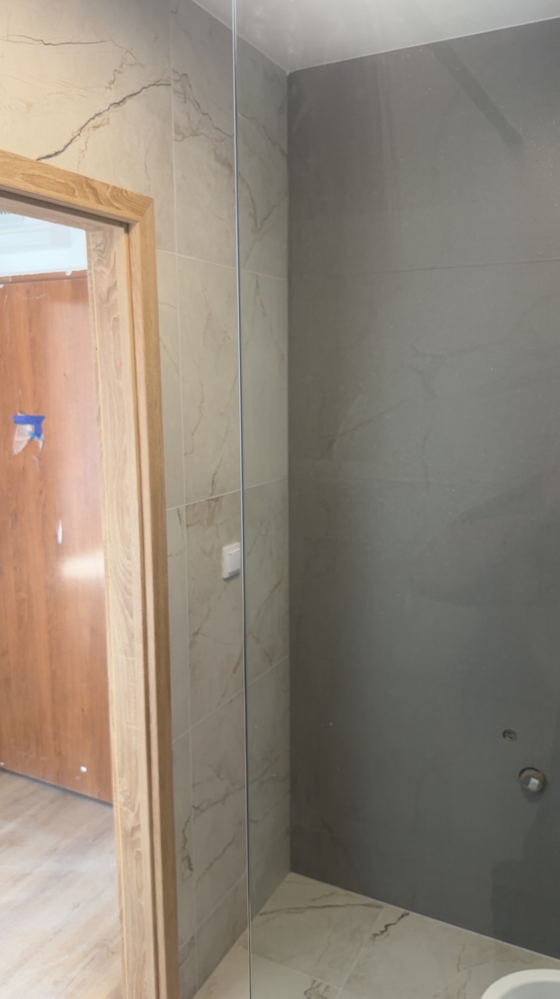Wykończona łazienka z beżowymi i szarymi płytkami imitującymi marmur, widoczna kabina prysznicowa i fragment drewnianej ościeżnicy drzwiowej.
