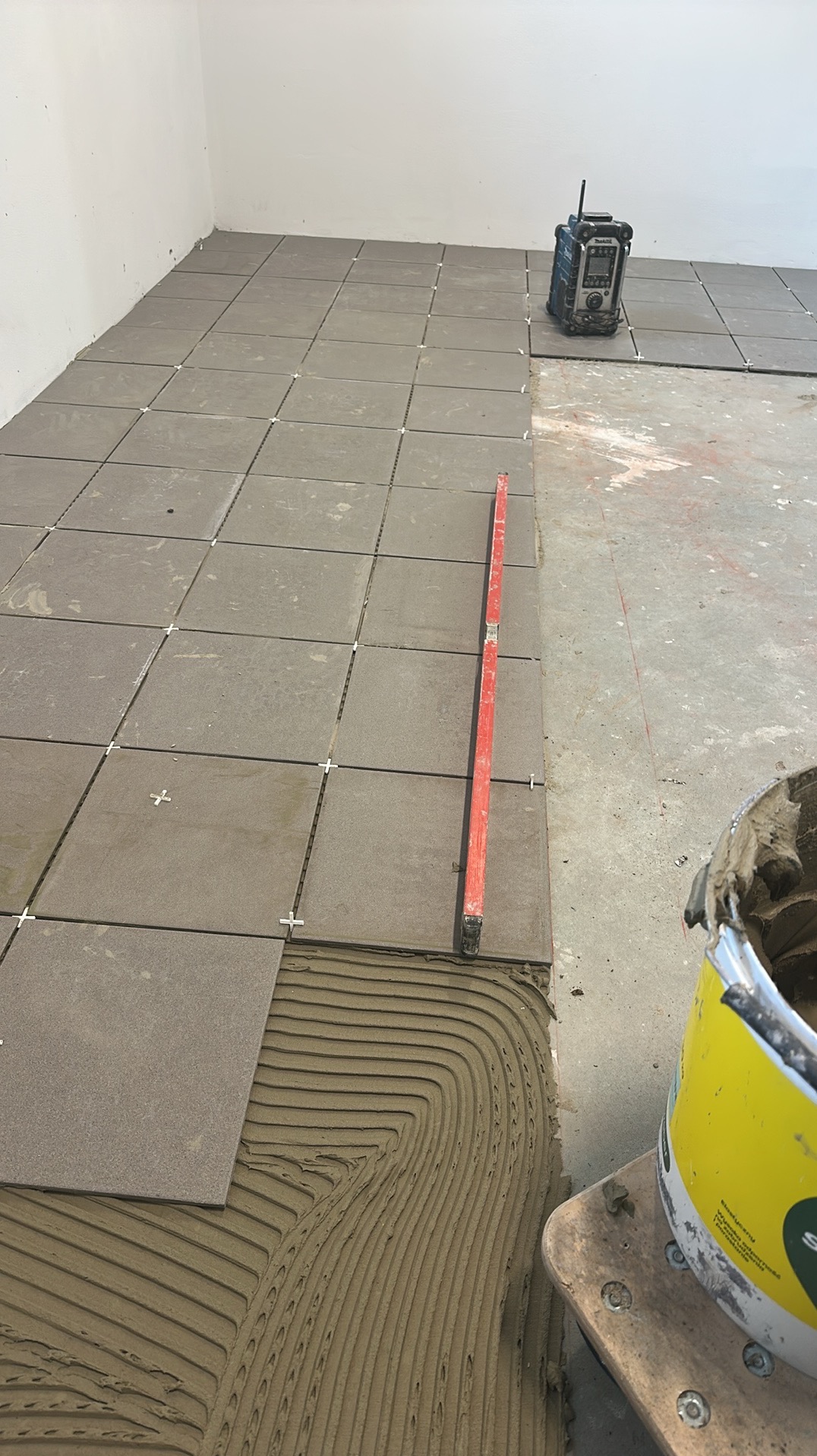 Układanie szarych, kwadratowych płytek podłogowych z użyciem poziomicy i krzyżyków dystansowych. Widoczna warstwa kleju i wiadro z zaprawą. Radio budowlane w tle.