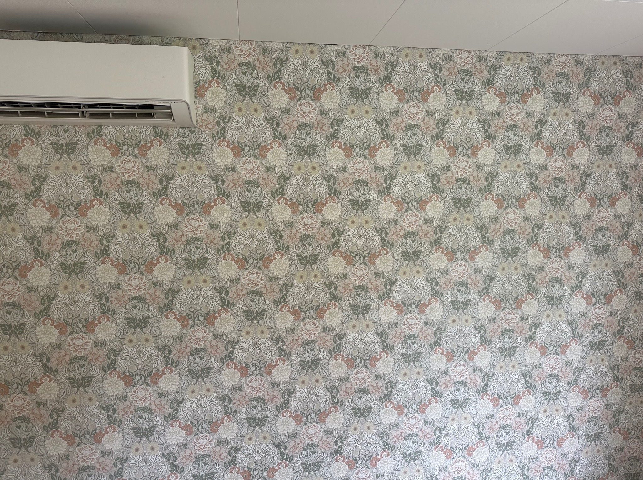 Ściana z tapetą w kwiaty i klimatyzator w Batorzu. Delikatny wzór w odcieniach szarości, różu i zieleni. Widoczny fragment sufitu. Subtelne wykończenie wnętrza.