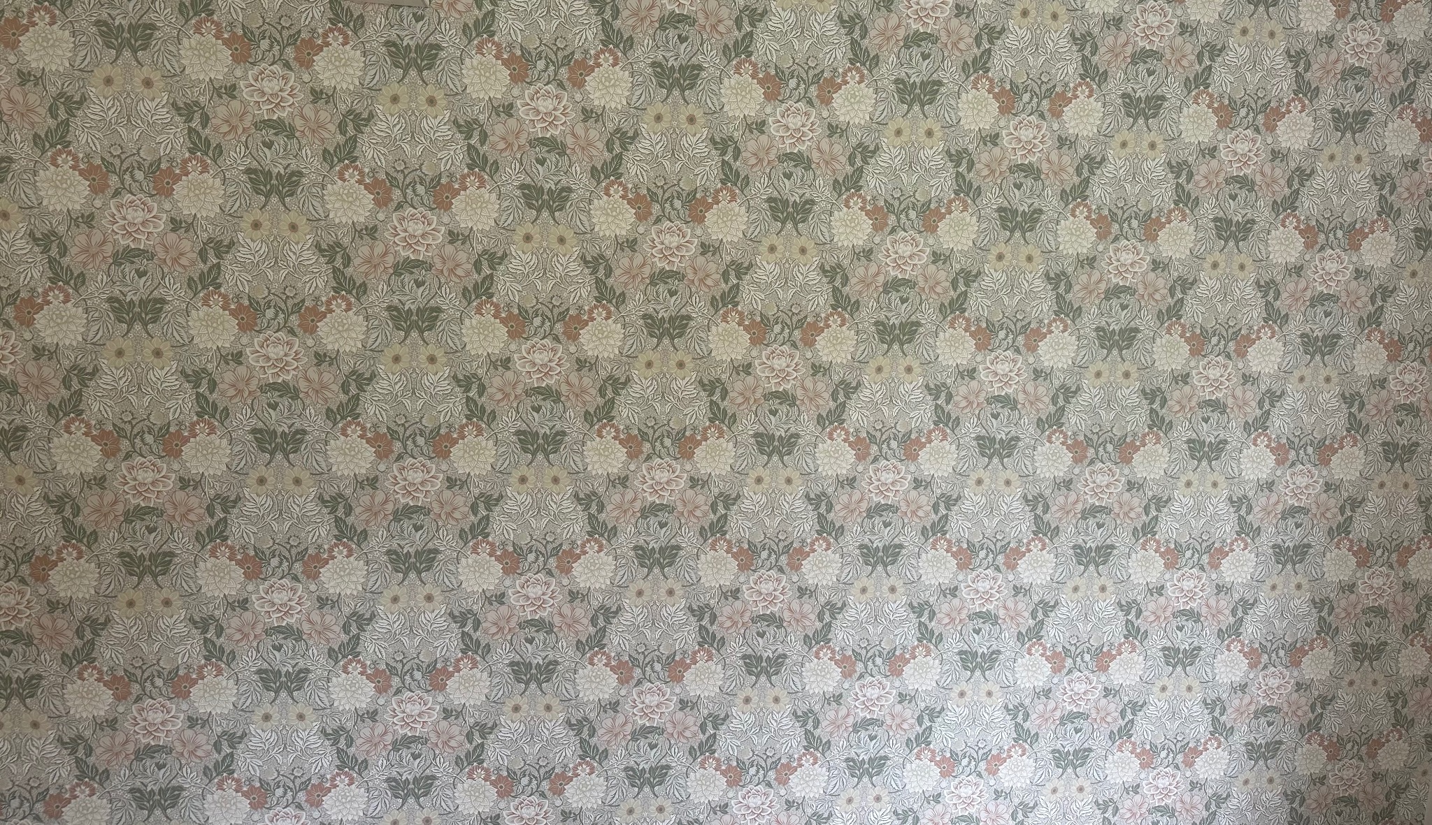 Ściana pokryta tapetą w stylu vintage z motywem kwiatowym w odcieniach beżu, różu i zieleni. Delikatny wzór, równomierne oświetlenie, brak widocznych łączeń tapety.