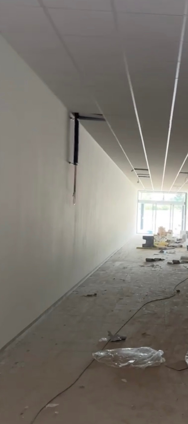 Długi korytarz biurowy w trakcie prac wykończeniowych: świeżo pomalowane ściany, sufit podwieszany z oświetleniem liniowym, widoczne narzędzia i materiały budowlane na podłodze.