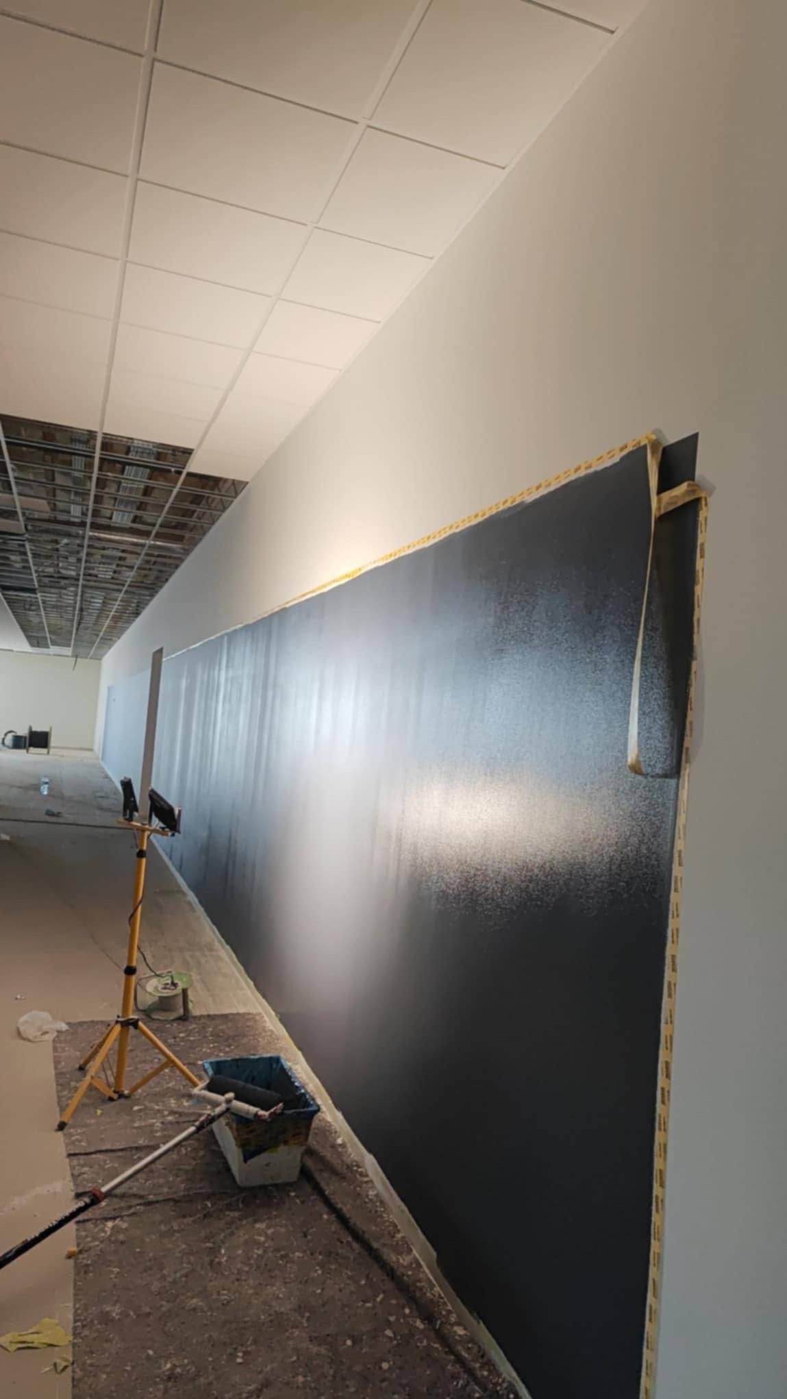 Ściana pomalowana na ciemny kolor w biurze, widoczne narzędzia malarskie, taśma malarska zabezpieczająca krawędzie, oświetlenie robocze. Prace wykończeniowe w toku.