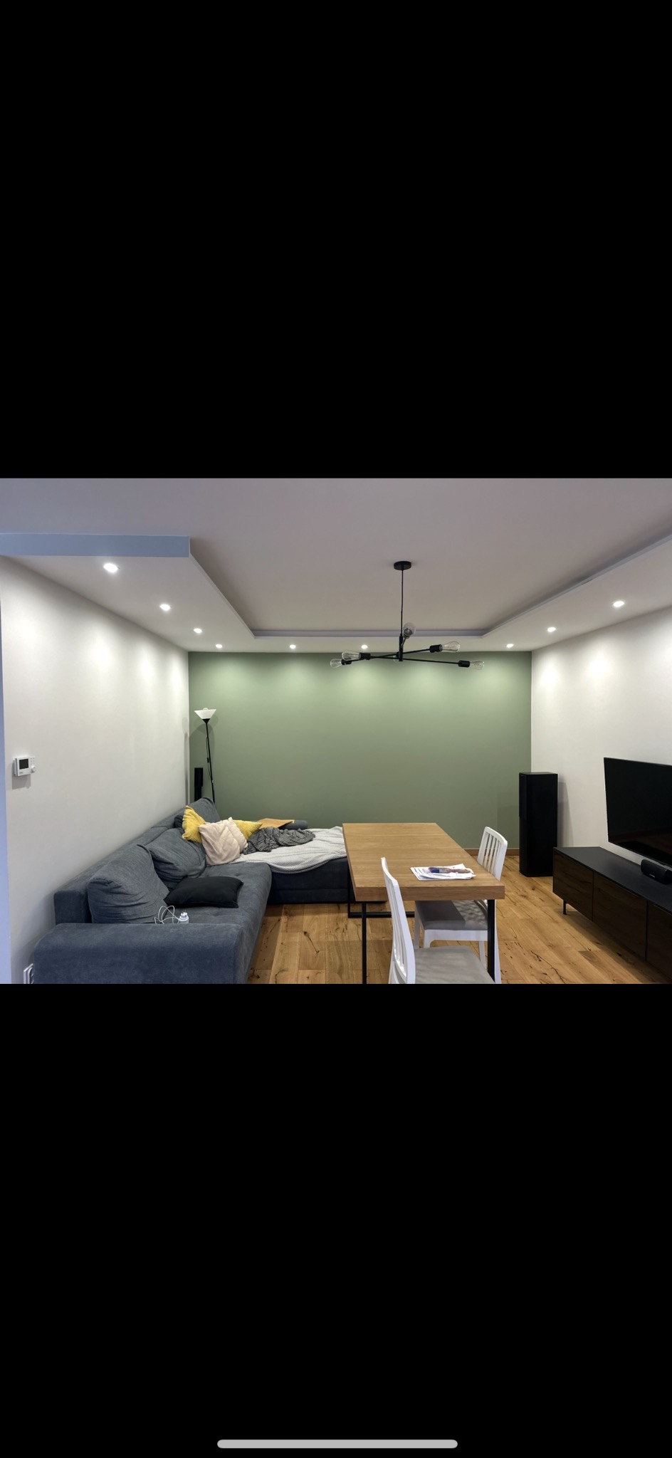 Salon po odświeżeniu: drewniana podłoga, szara sofa, stół z krzesłami, zielona ściana z oświetleniem punktowym i nowoczesna lampa sufitowa.
