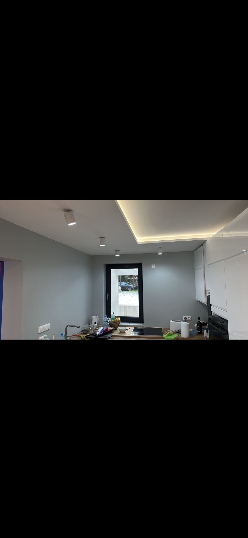 Jasna kuchnia po remoncie z szarymi ścianami, białymi szafkami i oświetleniem LED. Widok na okno z zielenią na zewnątrz. Minimalistyczny design.