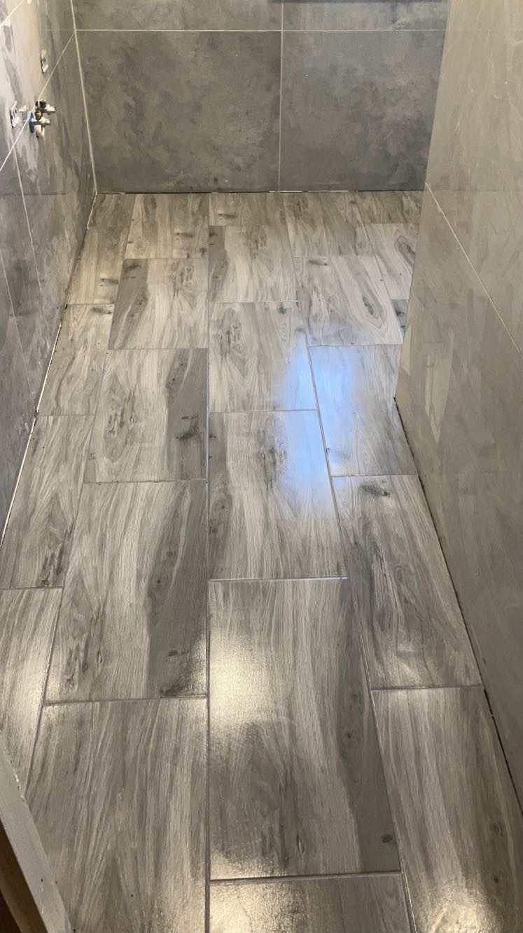 Ukończona łazienka z szarymi płytkami imitującymi drewno na podłodze i ścianach. Widoczne metalowe elementy instalacji wodnej.